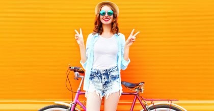 Eine junge Frau mit Sonnenbrille und Sonnenhut steht vor ihrem Fahrrad und zeigt das Peace-Zeichen.